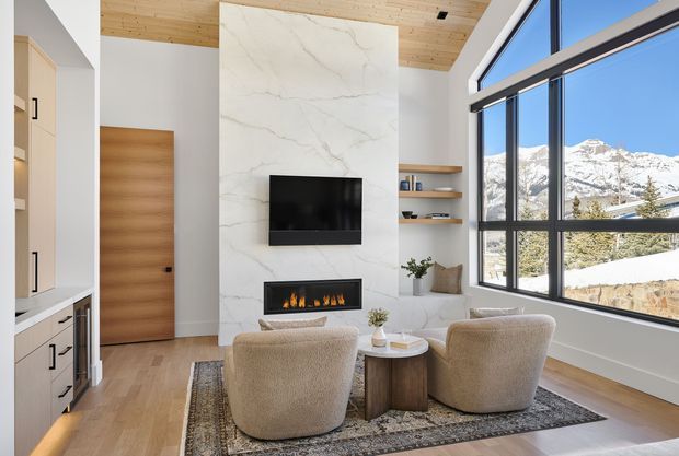 A bright contemporary living room with a double-height glazed wall overlooking snowy mountains. The room features a floor-to-ceiling veined stone fireplace and media wall, built-in shelving, a timber-clad ceiling, light timber flooring, a neutral sectional sofa, and two boucle lounge chairs around a small round coffee table.