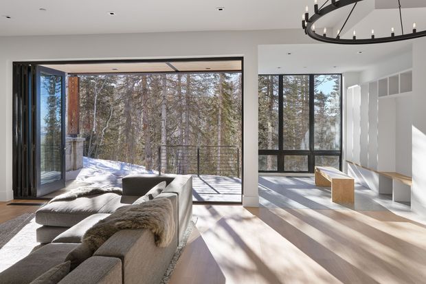 A bright, open-plan contemporary living room with extensive floor-to-ceiling glazing overlooking a wooded, snowy landscape. The room features a large modular grey sectional sofa, pale timber flooring, white walls and built-in cabinetry, black-framed windows and folding doors, and a prominent circular black metal chandelier. Sunlight creates strong shadows across the floor, with a deck visible beyond the open glazing.