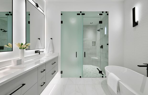 Bright contemporary bathroom with a long white double vanity, frameless glass walk-in shower, freestanding white bathtub, large mirrors, and white tiled surfaces. Black hardware and fixtures provide crisp contrast, while integrated lighting creates a clean, polished atmosphere.