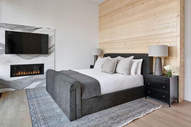 A contemporary bedroom with a large upholstered bed, layered neutral bedding, and a grey throw. The room features a full-height horizontal timber feature wall behind the bed, a black bedside table, matching lamps, pale timber flooring, and a large muted patterned rug. A marble-look fireplace wall with a wall-mounted television is visible to the left.