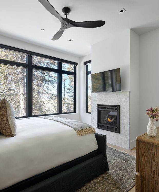 Bright, modern bedroom with a large upholstered bed positioned beside expansive black-framed corner windows. A white fireplace surround with a contemporary firebox and wall-mounted television forms the focal point, while a dark ceiling fan, neutral bedding, patterned rug, and timber bedside cabinet complete the calm interior.