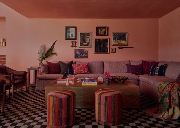 Eclectic living room with a large L-shaped upholstered sectional, patterned cushions, two striped ottomans, a broad wood-toned coffee table, and a black-and-cream checkerboard rug. The walls and ceiling are painted in warm terracotta-pink tones, with a gallery wall of framed artwork and a tall decorative plant adding colour and texture. The room has a cozy, richly layered, maximalist character.