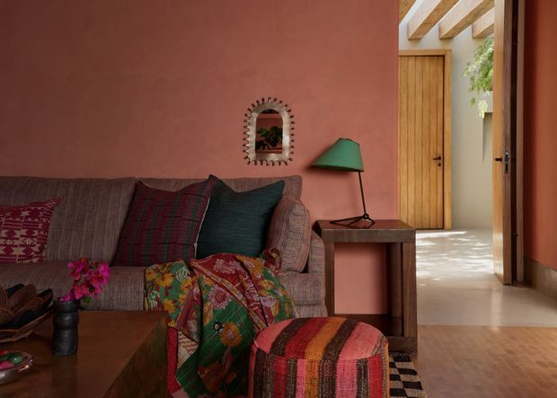 Warm, richly layered living room with a textured upholstered sofa, colourful patterned cushions and quilt, striped ottoman, dark wood coffee and side tables, and a green table lamp. Terracotta plaster walls continue into a bright adjoining passage with timber doors and exposed timber ceiling beams. An arched wall mirror and abundant textile pattern add an eclectic, handcrafted character.