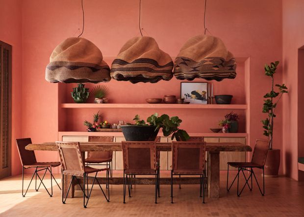 Warm, richly coloured dining room with a long rustic timber dining table and multiple woven leather or textile chairs. Three oversized sculptural woven pendant lights hang above the table. The coral-pink walls and built-in open shelving display ceramics, framed artwork, and potted plants, while a timber sideboard runs along the rear wall. Dark timber flooring and black metal chair frames add contrast.