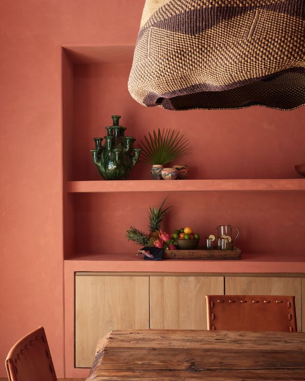 Warm dining area with terracotta plaster walls and built-in open shelving, featuring a woven pendant light, green ceramic vessels, decorative bowls, fruit, and a large rustic timber dining table. Light timber cabinetry occupies the lower portion of the shelving, with leather dining chairs partially visible.