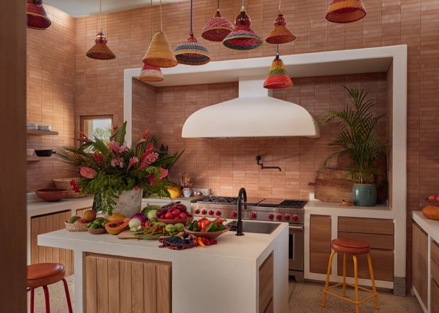 A warm, richly styled kitchen with a large central island, timber cabinetry, terracotta-toned rectangular wall tiles, and a prominent white sculptural rangehood. The island includes a dark benchtop section and integrated sink with a black tap. Colourful woven pendant lights hang from the ceiling, while open shelving, plants, flowers, produce, and stools add a lively decorative feel.