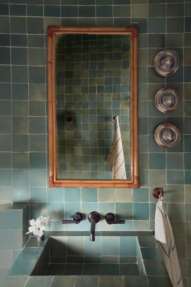 A compact bathroom vanity clad in dark teal-green square tiles, with a deep tiled basin, dark metal wall-mounted faucet, and a warm timber-framed rectangular mirror. Small round wall fixtures and hanging striped towels add detail, while soft natural light creates a calm, earthy atmosphere.