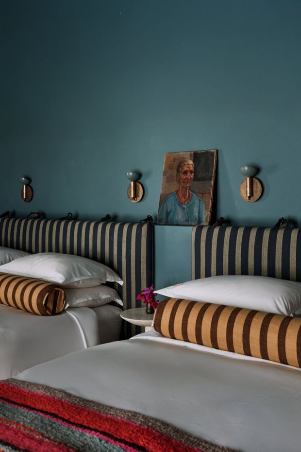 A moody, richly styled bedroom with two twin beds featuring tall navy-and-tan vertically striped upholstered headboards. The room has deep blue walls, warm brass and dark wall sconces, white bedding, striped cylindrical bolsters, a small round bedside table with flowers, and a framed portrait resting between the beds. A vivid red, charcoal, and grey woven throw adds contrast at the foot of the foreground bed.