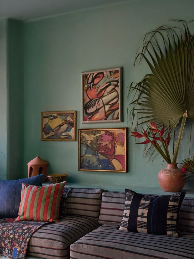 A richly styled living room with deep teal-green walls, a large striped sectional sofa, colourful patterned cushions, framed abstract and botanical artwork, and tropical foliage in decorative vessels. The layered textiles and saturated colours create an eclectic, warm, moody atmosphere.