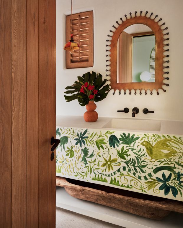 A compact, characterful bathroom viewed through a partially open timber door. The room features a white basin with matte black wall-mounted tapware, a large organic-shaped timber-framed mirror with decorative dark bead detailing, warm white walls, and a vanity front painted with a colourful botanical mural. A rustic timber shelf or vanity base sits beneath the basin, while a leafy arrangement and small wall artwork add natural, handcrafted character.