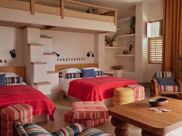 A colourful, warm bedroom with two beds, a loft-level sleeping or play area, and an open staircase with timber treads. The room features red bedspreads, striped upholstered chairs and ottomans, built-in shelving with plants and ceramics, timber furniture, woven balcony screening, and a timber-shuttered window. The overall design has a playful, richly patterned, resort-like character.
