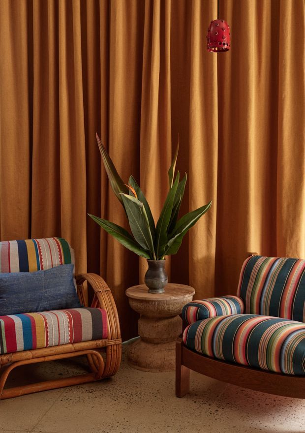 A richly styled seating area with two vintage-inspired armchairs upholstered in bold multicoloured stripes, positioned around a carved stone pedestal table holding a leafy tropical plant. Full-height pleated ochre-brown curtains form the backdrop, with warm natural light and a small red pendant or decorative hanging light above.
