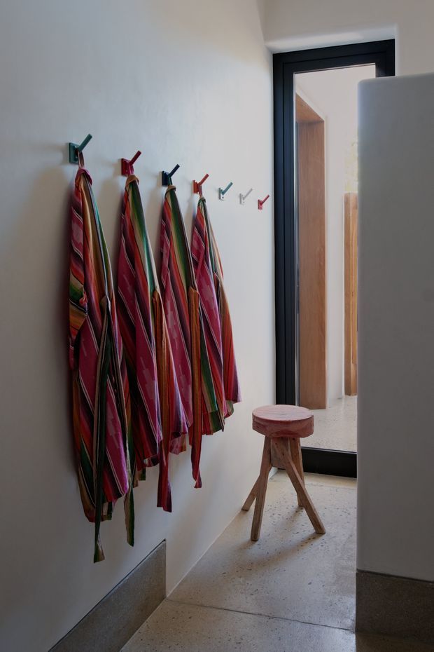 A narrow, bright entry or hallway with a white plaster wall lined with colourful striped fabric aprons or garments hanging from individual hooks. A small rustic wooden stool sits on a polished speckled concrete floor near a dark-framed glazed door. The space has a simple contemporary fit-out with strong textile colour accents and natural timber visible beyond the doorway.