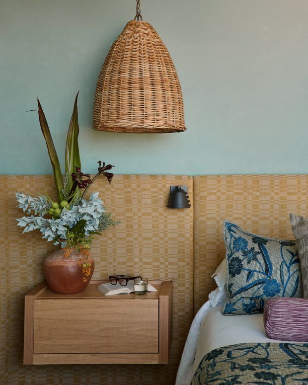 A warmly styled bedroom corner with a low timber bedside cabinet beside a bed. The room features a blue-green painted upper wall, a woven tan patterned wall covering or tile-like finish below, a large woven rattan pendant light, botanical arrangement in a terracotta vessel, and layered blue-and-white patterned bedding and cushions. The composition feels eclectic, earthy, and inviting.