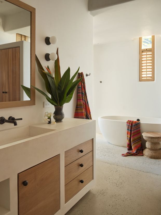 Warm, refined bathroom interior featuring a sculptural white freestanding bathtub, a floating or partially built-in timber vanity with pale stone or plaster-like benchtop, wall-mounted dark tapware, a large timber-framed mirror, and a small timber shuttered window. A colorful striped textile and tropical plant add natural and vibrant accents. The space has a light textured floor, white walls, and a calm contemporary feel.