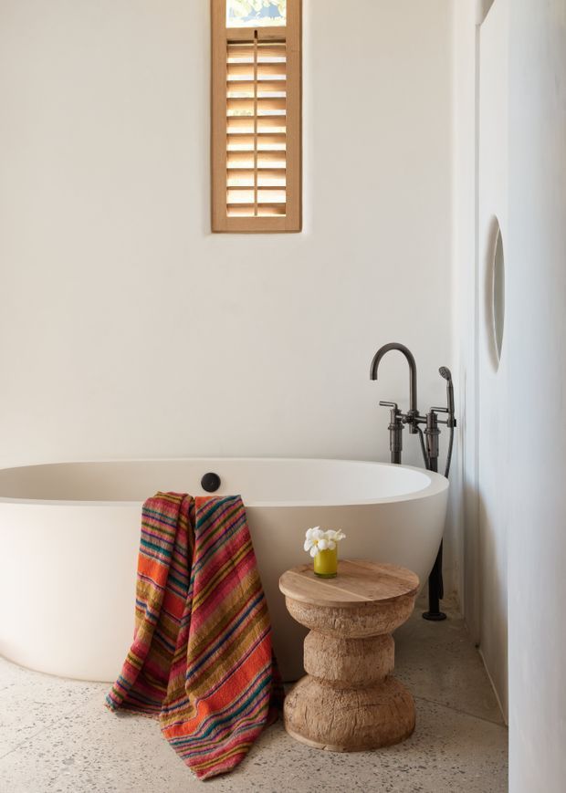 A bright, calm bathroom featuring a freestanding oval white bathtub, dark metal floor-mounted tub filler, and a small sculptural natural timber side table. A colorful striped towel drapes over the bath, while a narrow timber louvred window adds warm texture above. The room has white walls and a speckled concrete or terrazzo-style floor.