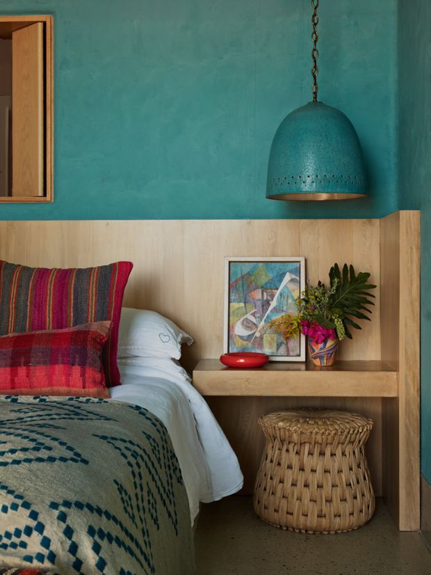 A richly styled bedroom corner with a built-in timber headboard and bedside shelf, turquoise textured wall, colorful woven bedding and cushions, a woven basket stool, framed artwork, and a large turquoise pendant light. The space has a warm, handcrafted, eclectic character with strong jewel-toned colour accents.