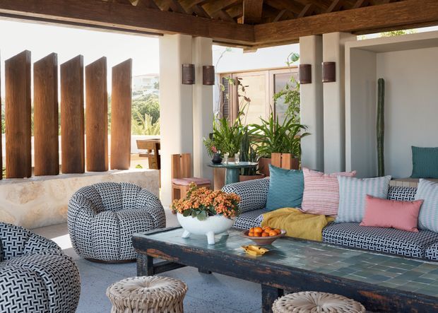 A bright, open-air living lounge with exposed timber beams and rafters, white rendered columns and walls, patterned upholstered armchairs and sofa, colourful cushions, abundant potted greenery, and a long dark timber coffee table with a tiled top. Vertical timber screening provides privacy at the open edge, creating a relaxed indoor-outdoor entertaining space.