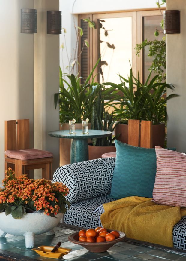 A warmly styled indoor conservatory or sunroom with abundant potted greenery, a wood-framed glazed opening, sculptural wall lights, and a patterned lounge chair. The space includes a small round pedestal table, timber stools, colourful cushions, a mustard throw, and a tiled coffee table with decorative objects and a bowl of fruit. The overall look is richly layered, plant-filled, and eclectic with strong mid-century influences.