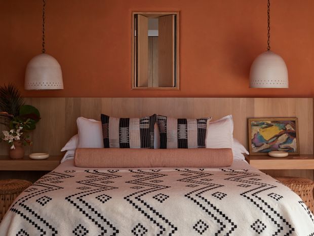 Warm, richly styled bedroom with a low platform bed against a timber-panelled headboard wall. The room features terracotta-painted walls, a geometric black-and-cream bedspread, layered patterned cushions, woven bedside furniture, paired oversized pendant lights, a central timber-framed mirror, and organic decorative accents.