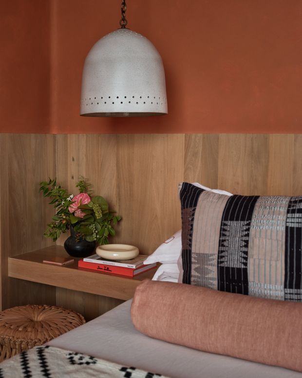 Close-up of a warm, richly styled bedroom corner featuring a bed with patterned black, cream and grey cushions, a long blush-pink bolster, and neutral bedding. A timber wall and built-in bedside shelf sit behind the bed, decorated with books, a ceramic bowl and a small floral arrangement. A large perforated pendant light hangs against a terracotta painted wall, creating a cosy, earthy atmosphere.