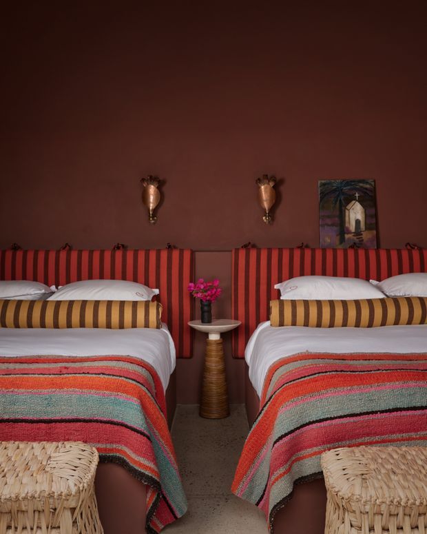 A richly styled bedroom with two twin beds separated by a narrow pedestal table. The beds feature deep red striped upholstered headboards, layered white bedding, multicoloured woven blankets, and striped bolster cushions. The room has warm dark brown walls, decorative brass-toned sconces, a small religious artwork, woven bedside furniture, and a textured neutral floor. The overall atmosphere is bold, warm, eclectic, and intimate.