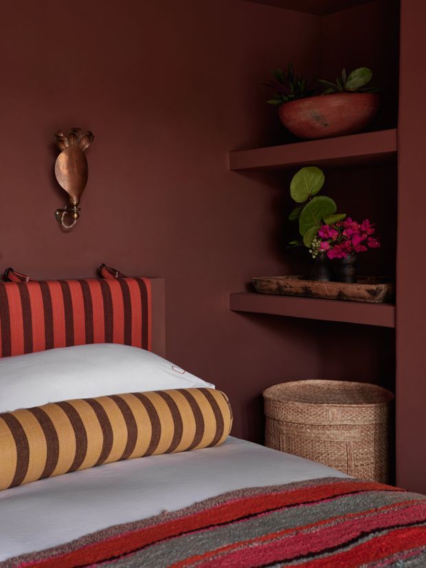 A richly coloured, moody bedroom corner featuring a bed with bold red, brown and mustard striped upholstery and bedding. Deep burgundy painted walls frame built-in open shelves displaying potted plants and decorative objects, with a woven basket beside the bed and a small ornate wall light. The styling is warm, layered and eclectic with strong pattern and texture.
