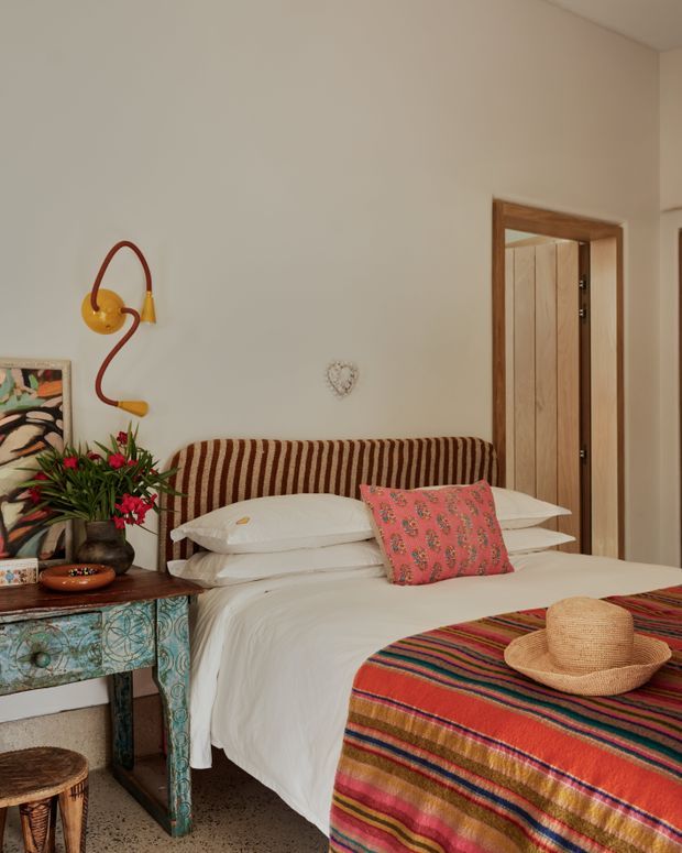 A warmly styled bedroom with a white bed layered in pillows, a striped upholstered headboard, and a vibrant multicoloured woven throw across the foot. A patterned painted bedside table holds flowers and decor, while a sculptural wall lamp, framed artwork, and timber doorway add character. The room has light neutral walls and a terrazzo-like floor, with eclectic global-inspired furnishings and textiles.