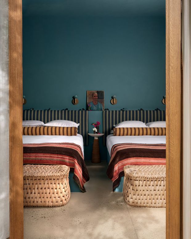 A compact twin bedroom viewed through a timber-framed doorway, with two single beds arranged symmetrically against a deep teal wall. The beds feature striped upholstered headboards, layered striped and woven blankets, white bedding, and woven rattan footstools. A small central bedside table with flowers and wall-mounted lights completes the warm, eclectic composition.
