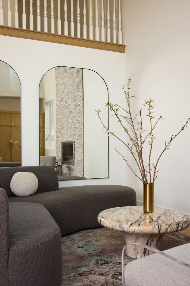 A refined living room with a large dark charcoal sectional sofa, sculptural marble coffee table, patterned area rug, and a gold vase with bare branches. Oversized arched mirrors reflect a pale stone fireplace and timber cabinetry, while a traditional timber-trimmed balustrade overlooks the room above. The palette is warm neutral with a sophisticated contemporary mix of classic architectural detailing and modern furnishings.