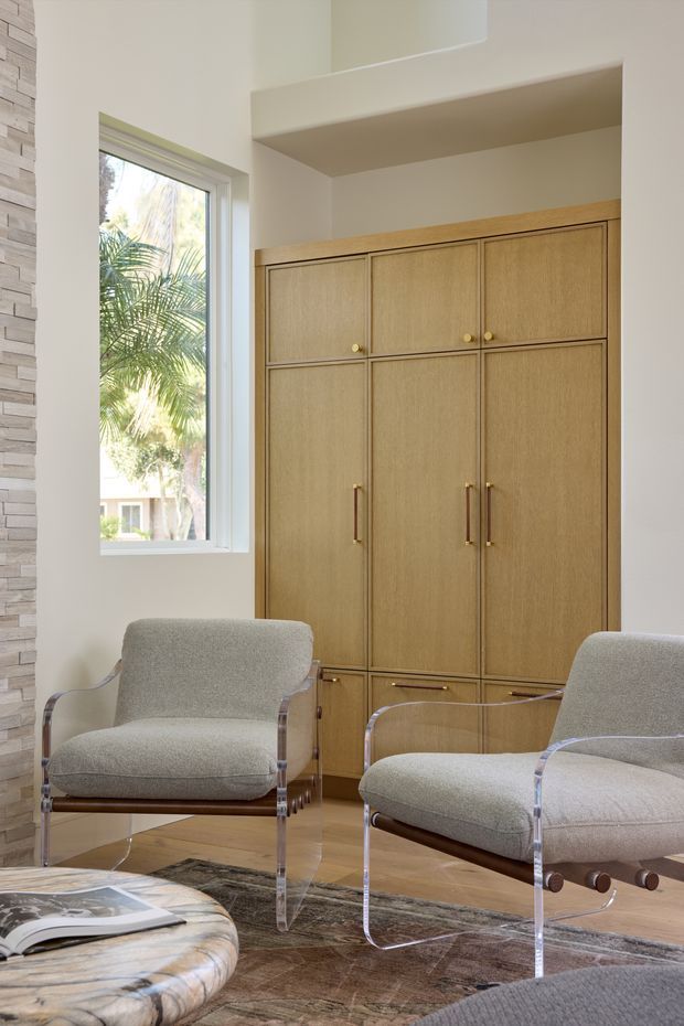 Bright, refined interior seating area with two upholstered lounge chairs featuring clear acrylic frames and dark wood supports. A large built-in timber cabinet with brass hardware occupies the wall, beside a tall window overlooking tropical greenery. Pale walls, timber flooring, a sculptural stone coffee table, and a patterned area rug create a warm contemporary setting.