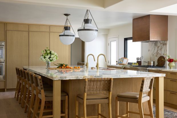 A bright, spacious contemporary kitchen with a large marble island and integrated seating for several people. The room features light timber cabinetry, fluted and woven detailing on the island base and stools, brass-toned tapware, pendant lights, and a veined stone splashback. Full-height cabinetry and integrated appliances line the walls, creating a refined, warm, natural interior.