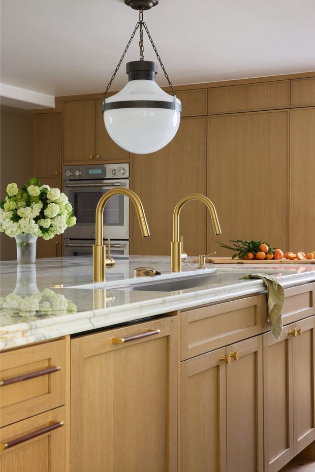 A warm, refined kitchen with extensive light-to-mid timber cabinetry, a large pale marble island benchtop, twin curved brass-gold gooseneck faucets, integrated stainless-steel appliances, and a large vintage-inspired pendant light. Fresh flowers and citrus add decorative colour, while the cabinetry and island dominate the composition.