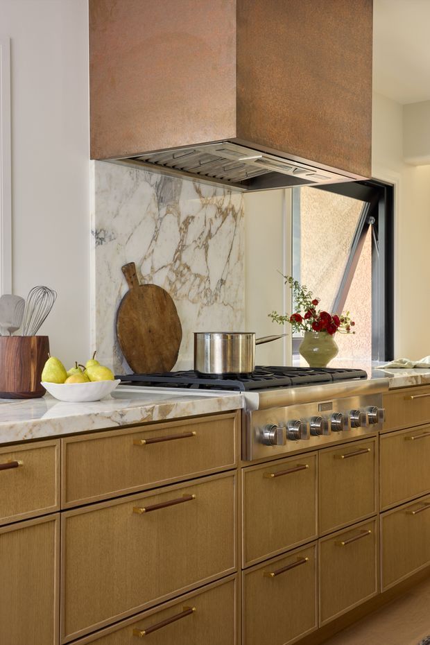 Close-up view of a refined kitchen featuring warm wood cabinetry with brass-and-wood pulls, a large professional gas range, white marble countertops and backsplash with prominent veining, and a substantial copper-toned metal range hood. A dark-framed window provides natural light, while simple countertop accessories and a vase of flowers add warmth.
