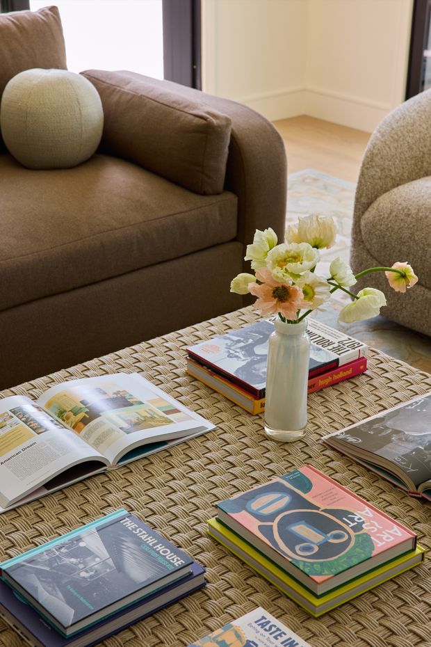 Close-up interior view of a warm, refined living room vignette. A brown upholstered sofa and textured rounded armchair surround a large woven coffee table topped with stacked design books, open magazines, and a simple vase of pale flowers. Soft neutral walls, light timber flooring, and natural textures create a calm, inviting atmosphere.