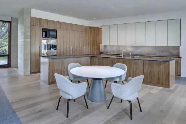 A bright, open-plan contemporary kitchen and dining area with extensive timber cabinetry, a large island, pale upper cabinets, integrated appliances, and a round dining table with four upholstered chairs. The room features light timber flooring, a pale stone or tile backsplash, white walls and ceiling, and broad glazing along the left side.