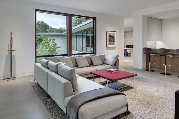 Contemporary open-plan living room with a large pale sectional sofa arranged around a low red coffee table. The room features light neutral walls, a broad textured rug, dark timber-framed glazing, and a partially visible kitchen or bar area with timber cabinetry and stools. A landscaped courtyard and adjacent modern structure are visible through the windows.