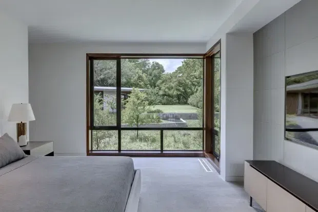 A bright, minimalist bedroom with a low-profile upholstered bed, neutral grey bedding, a simple bedside table and lamp, and a large timber-framed corner window overlooking dense greenery and a neighbouring modern structure. A wall-mounted television and streamlined low cabinetry sit along the right wall. The room has a calm, contemporary feel with pale carpet and white walls.