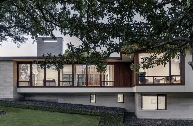Exterior view of a contemporary, low-rise residential house set behind mature trees. The facade combines white rendered walls, dark grey masonry at the lower level, extensive black-framed glazing, and warm timber vertical screening and cladding. A flat roofline and a raised upper volume create a clean horizontal composition, with landscaped lawn and planting in the foreground.