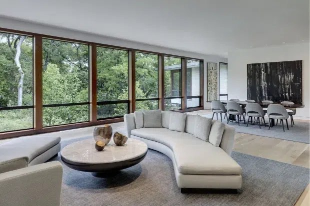 A spacious contemporary open-plan living and dining area with a large curved sectional sofa, rounded coffee table, and neutral upholstered seating. Full-height timber-framed glazing runs along one side, opening views to dense greenery. The dining zone features a long table, multiple grey chairs, and a large abstract dark artwork. Neutral carpeting and light timber flooring create a calm, refined interior.