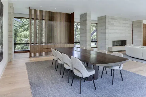 Open-plan contemporary dining and living area with a long dark timber dining table surrounded by upholstered chairs, set on a large grey rug over pale timber flooring. White stone-clad structural columns and fireplace, extensive black-framed glazing, and a vertical timber slatted screen create architectural definition while connecting the room to leafy outdoor views.