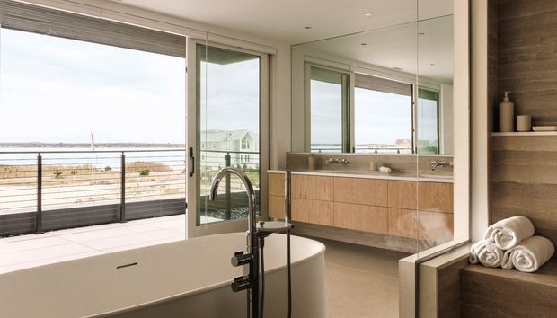 Bright contemporary bathroom with a freestanding white bathtub, wall-mounted timber vanity, large mirror, and open shelving with rolled towels. Full-height glazing opens to a terrace and provides a broad coastal or waterfront view. The space has clean lines, light neutral finishes, and abundant natural light.