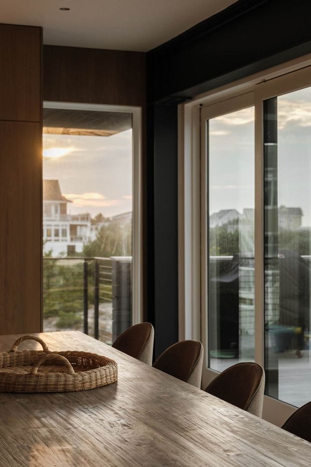A warm, moody dining area centered on a long natural timber dining table with curved brown upholstered chairs and a woven basket centerpiece. Dark timber wall cabinetry and black-framed structural elements contrast with large glazed doors and windows opening onto a balcony or outdoor area. The view beyond shows neighbouring coastal-style houses and soft evening light.