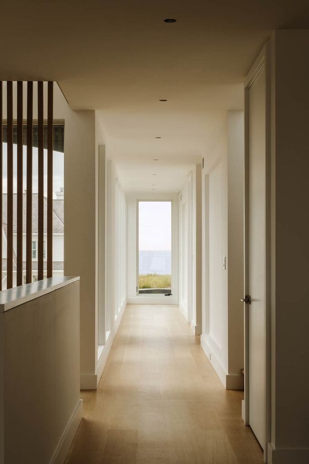 A long, bright residential hallway with pale timber flooring, white walls, recessed ceiling lights, and multiple white doorways. A partially open door is visible in the foreground, while a large glazed opening at the far end frames a distant coastal view. Vertical dark timber screening and a half-height built-in partition appear to the left.