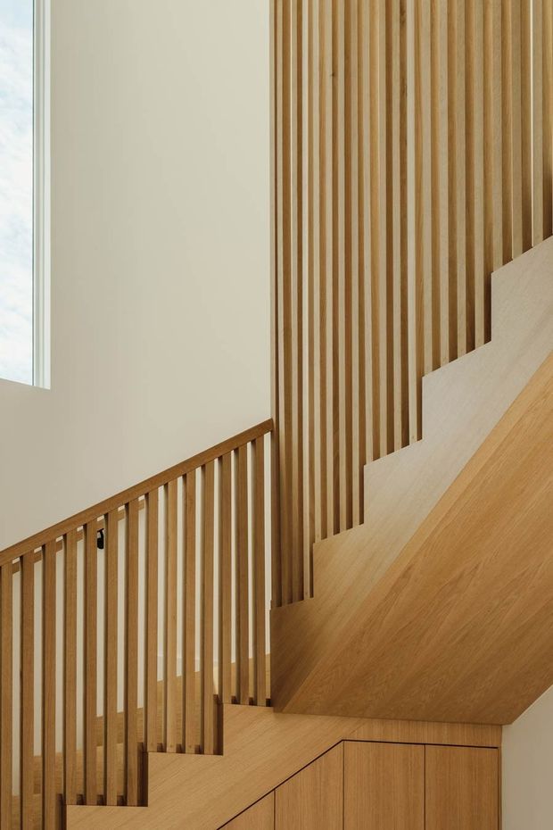 A close-up architectural view of a refined timber staircase and balustrade. Warm, light-toned wood treads and integrated joinery are paired with slender vertical timber slats extending up the wall, creating a continuous screen-like feature. A tall window brings soft natural light into the stairwell, while the clean geometry and minimal detailing give the space a contemporary, calm character.