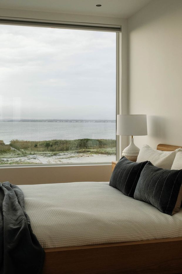 A calm, minimalist bedroom with a platform bed positioned beside a large picture window overlooking a sandy shoreline, water, and a distant coast. The room has off-white walls, a warm timber bed base, layered cream and white bedding, dark charcoal cushions and throw, and a simple white ceramic table lamp. The window has a slim metallic frame and roller blind, creating a quiet coastal outlook.