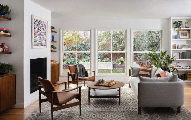 Bright, refined living room with three large traditional-style windows overlooking a leafy garden. The room features a grey sofa, mid-century leather lounge chairs, a round wooden coffee table, patterned area rug, built-in shelving, timber cabinetry, indoor plants, artwork, and a minimalist black fireplace set into a white wall.
