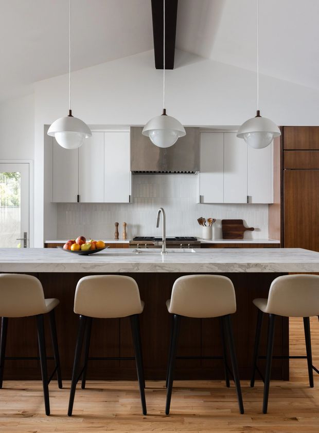 A refined contemporary kitchen viewed straight on, featuring a large dark timber island with a pale stone benchtop and four upholstered bar stools. The kitchen has white flat-panel cabinetry, a light vertical tile splashback, integrated stainless-steel appliances, warm timber flooring, and three suspended globe pendant lights. A tall timber storage wall adds contrast at the right.