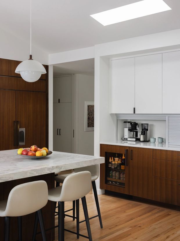 A bright, refined contemporary kitchen with a large marble-look island and integrated seating. The room features rich dark timber cabinetry, white upper cabinets, a concealed coffee station, built-in bar storage, warm timber flooring, and a sculptural white pendant light. A skylight brings additional natural light into the space.