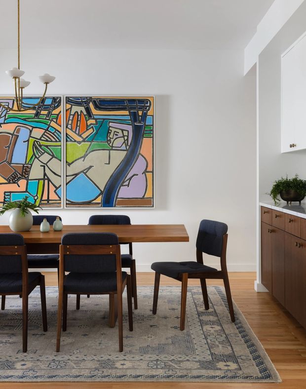 A bright, refined dining room with a mid-century-style timber dining table and upholstered dark chairs arranged on a large patterned rug. Two colourful abstract artworks hang on the white wall, accompanied by a brass-toned multi-arm pendant light. A built-in timber sideboard with a light stone countertop occupies the right side, with a tall opening leading toward an adjacent space. Timber flooring and clean white walls create a warm contemporary setting.
